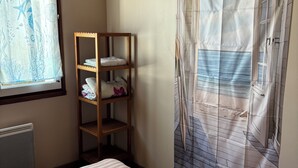 Et soveværelse med en seng, en træhylde med sammenfoldede håndklæder, en radiator og et vindue med gardin.