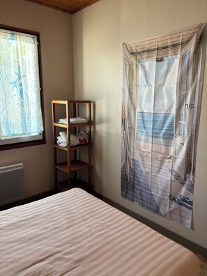 A bedroom with a bed, a wooden shelf with folded towels, a radiator, and a window with a curtain. - Seaside (La Tranche-sur-Mer)