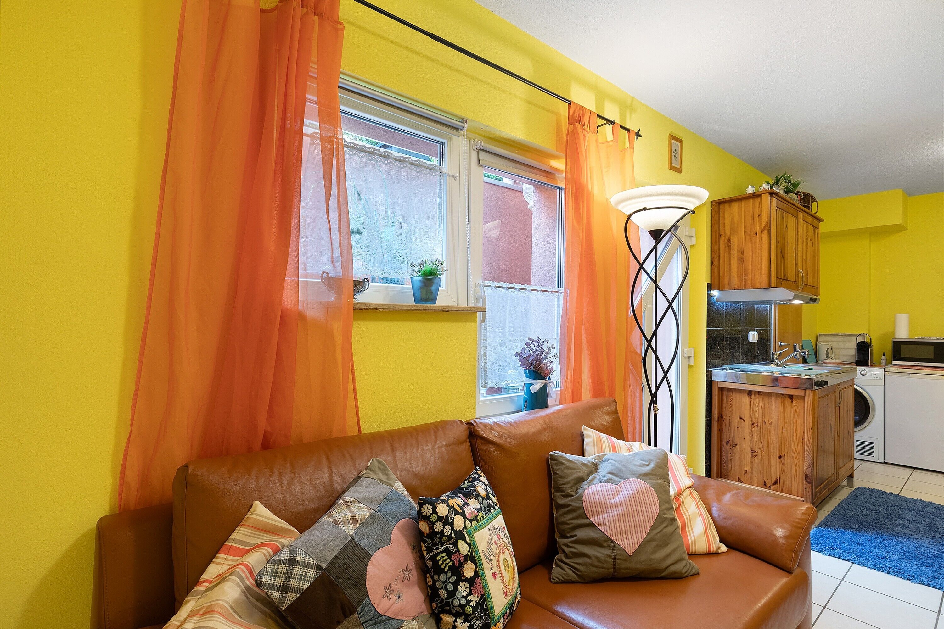 A living room with a brown leather sofa, orange curtains, a wooden cabinet, and a kitchen area with a washing machine.