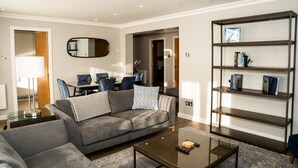 Un salon moderne avec un canapé gris, une table basse en verre, une bibliothèque et un coin repas comprenant des chaises et une table.