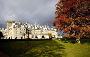 A large, historic building with multiple windows and a symmetrical facade, surrounded by a well-maintained lawn and a tree with autumn foliage.