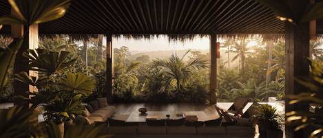 A covered outdoor seating area with a sofa and chairs, surrounded by tropical plants and a view of a forested landscape.
