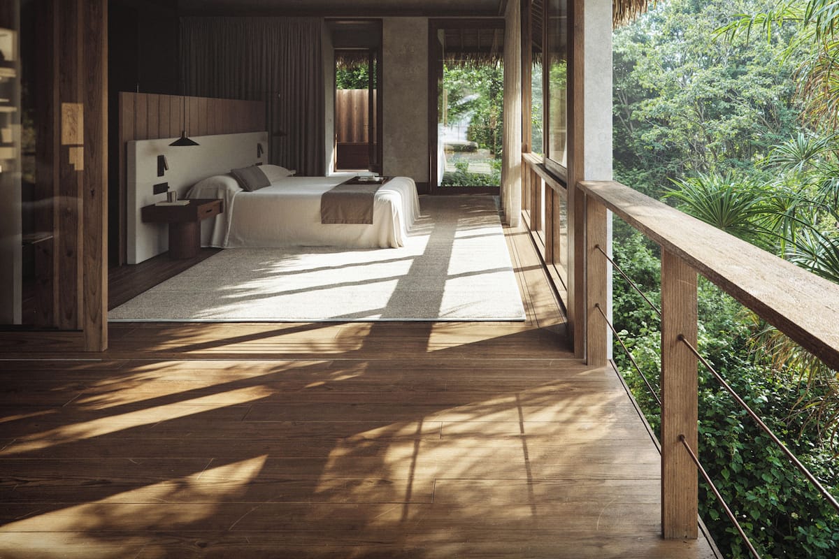 Un dormitorio amplio con una cama grande, una mesita de noche y un ventilador de techo. Cuenta con un balcón con baranda de madera que da a un jardín exuberante.