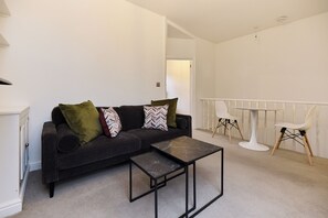 Ein Wohnzimmer mit einem dunklen Sofa, zwei gemusterten Kissen, einem kleinen schwarzen Couchtisch, einem weißen Tisch mit Stühlen und einer weißen Geländer.