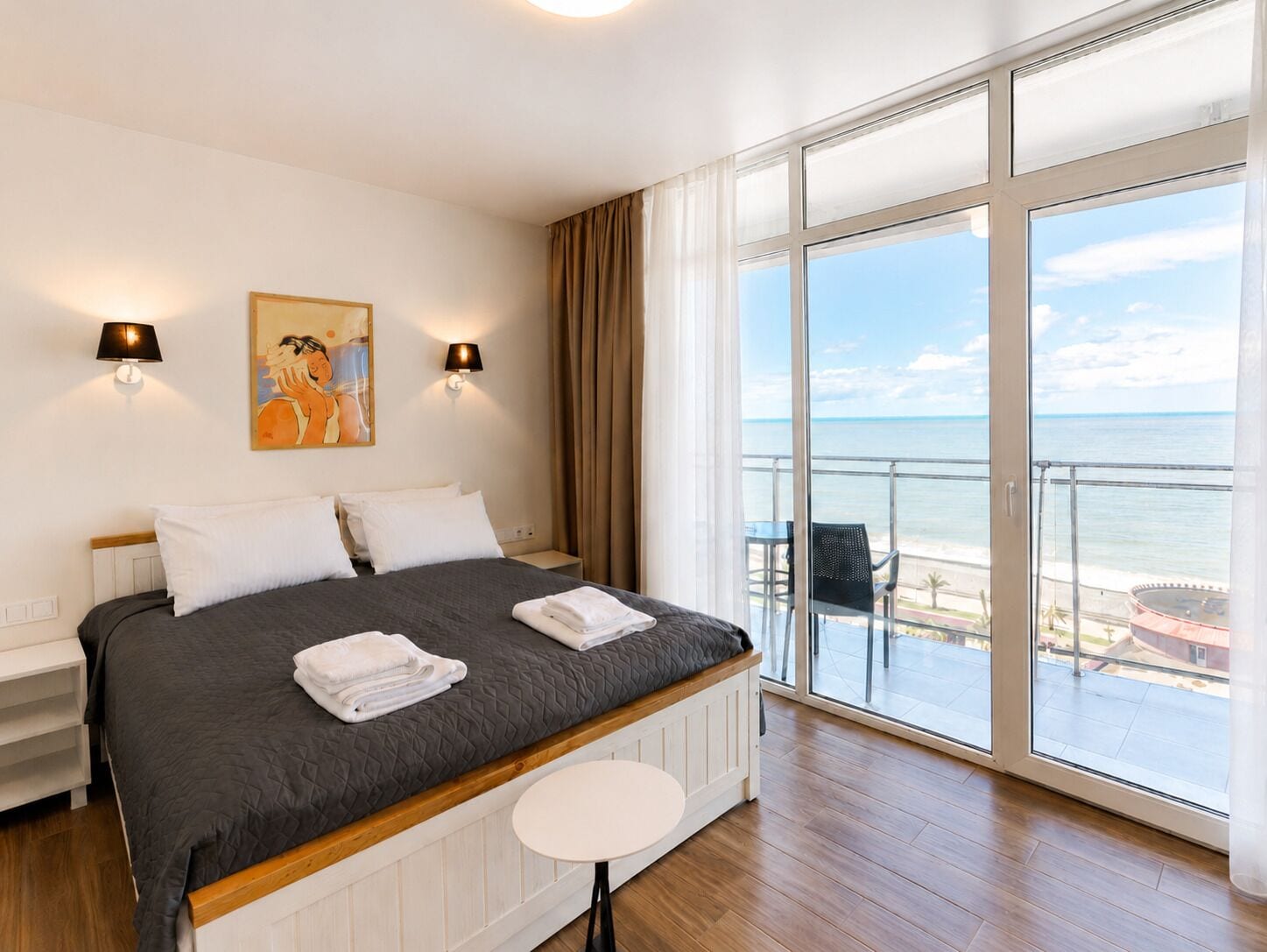 A hotel room with a bed, two wall-mounted lamps, a painting, a balcony with a view of the sea, and a small table.