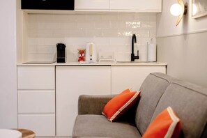A compact kitchen with white cabinets, a black faucet, and a grey sofa with orange cushions. - Babylon Popincourt 12 (Paris)