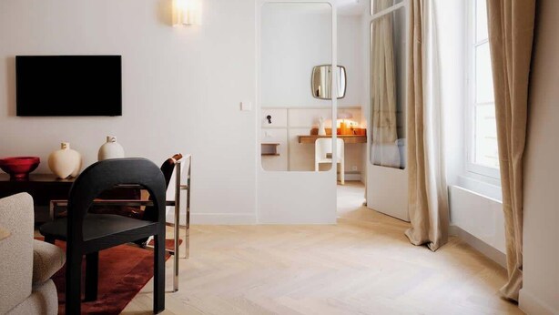 Ein modernes Wohnzimmer mit Flachbildfernseher, Essbereich mit Tisch und Stühlen, Küchenzeile mit Arbeitsfläche und Schränken sowie ein großes Fenster mit Vorhängen.