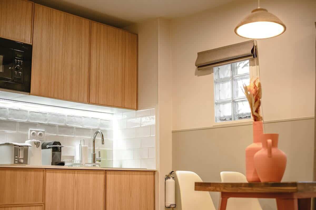 Una cocina con armarios de madera, una encimera blanca, una cafetera, un lavabo y un ventanal con cortina.