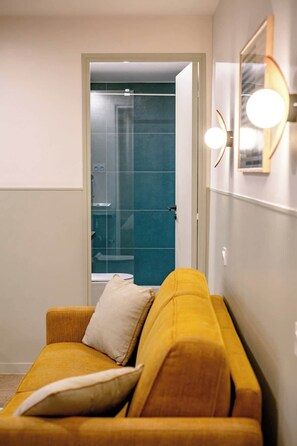 Ein Zimmer mit einem gelben Sofa, einem weißen Kissen, einer Glastür zum Badezimmer und einer wandmontierten Lampe.