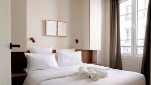 Ein Hotelzimmer mit einem ordentlich bezogenen Bett, einem Kopfteil, einem an der Wand hängenden Bild und einem mit Vorhängen versehenen Fenster.
