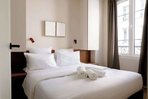 Ein Hotelzimmer mit einem ordentlich bezogenen Bett, einem Kopfteil, einem an der Wand hängenden Bild und einem mit Vorhängen versehenen Fenster.