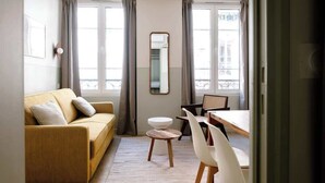Ein modernes Hotelzimmer mit einer Couch, einem kleinen runden Tisch, einem Stuhl, einem runden Hocker, einem Spiegel und einem Fenster mit Vorhängen.