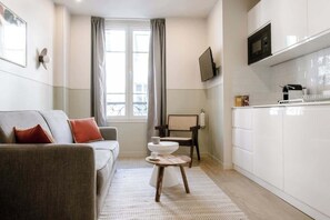 Ein modernes Wohnzimmer mit einer Couch, einem kleinen runden Tisch und einer Küchenzeile mit weißen Schränken und einer Mikrowelle.