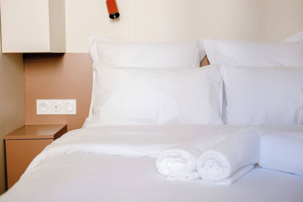 Una cama bien hecha con sábanas blancas y toallas enrolladas, una mesita de noche marrón con toma de corriente y una lámpara de pared.