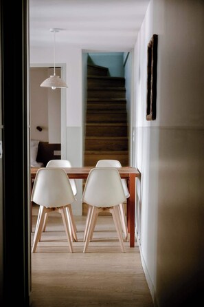 A dining area with a wooden table and white chairs, a hanging light fixture, and a staircase in the background. - Babylon Popincourt 23 (Paris)