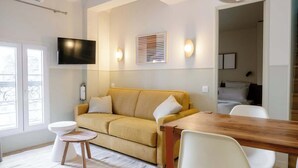 Een moderne woonkamer met een bank, een houten eettafel, een aan de muur bevestigde televisie en een raam met jaloezieën.