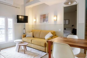 A modern living room with a sofa, a wooden dining table, a TV mounted on the wall, and a window with blinds. - Babylon Popincourt 23 (Paris)