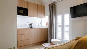 Een moderne keuken met houten kasten, een wit aanrechtblad en een ingebouwde magnetron. Er staat een klein rond tafeltje met een witte stoel, en een flatscreen televisie hangt aan de muur.