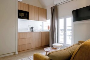 A modern kitchen with wooden cabinets, a white countertop, and a built-in microwave. There is a small round table with a white chair, and a flat-screen TV mounted on the wall. - Babylon Popincourt 23 (Paris)