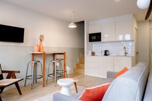 Eine moderne Küche mit weißen Schränken, einer Mikrowelle und einer Kaffeemaschine. Ein Essbereich mit Holztisch und zwei Hocker. Im Wohnzimmer steht ein graues Sofa mit orangefarbenen Kissen.