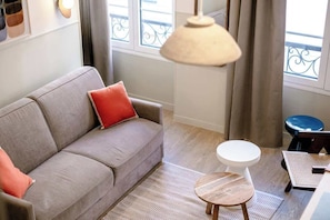 Ein Wohnzimmer mit grauem Sofa, einem roten Kissen, einem runden Holztisch, einer weißen Toilette und einem Balkon mit Metallgeländer.