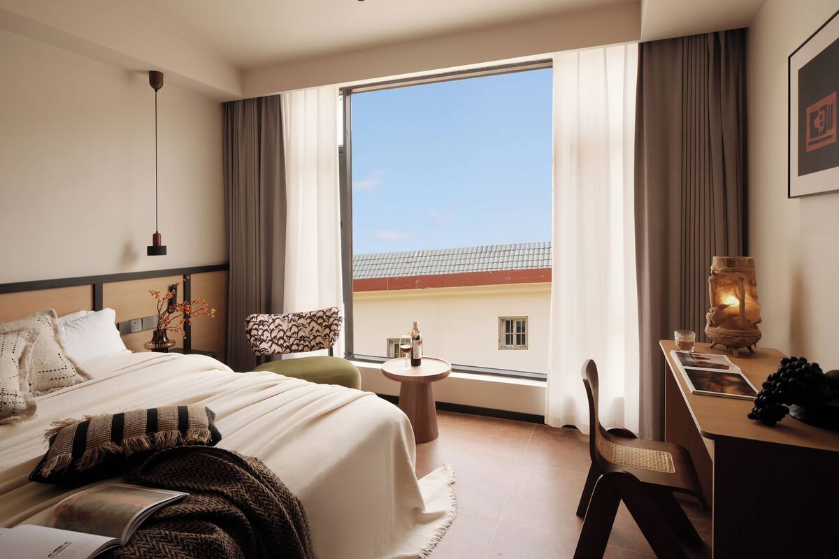 Habitación de hotel con cama, escritorio, silla, mesita pequeña y un amplio ventanal con vistas a edificios.