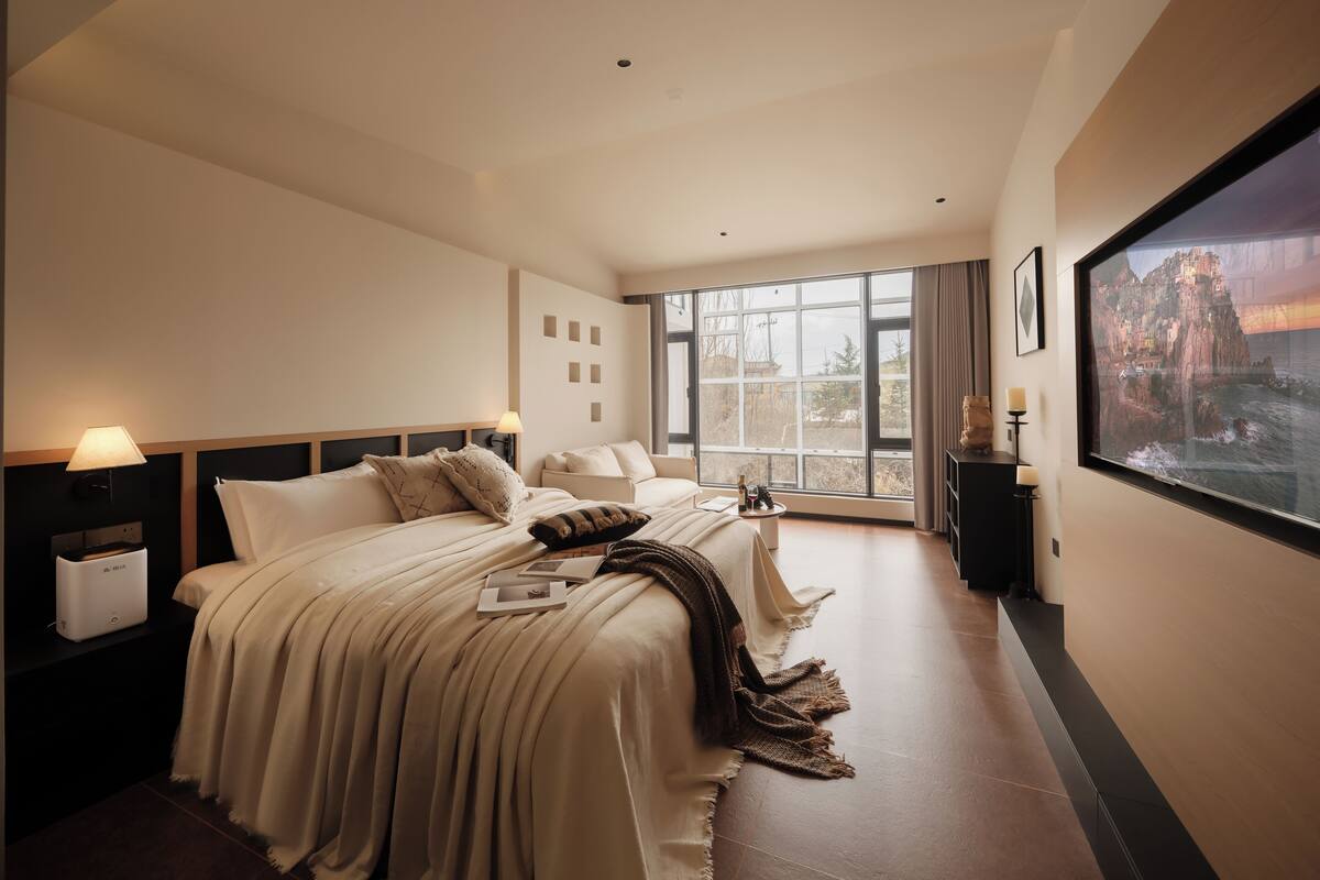 Habitación de hotel con una cama grande, una cama más pequeña, una ventana con vistas, una mesita de noche y un televisor montado en la pared.