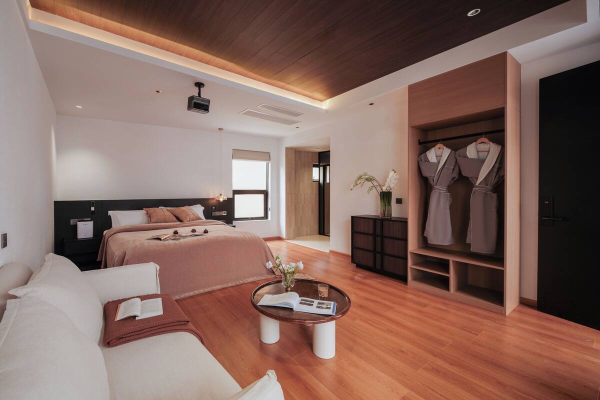 Un dormitorio moderno con una cama grande, un sofá, una mesita de centro con un libro y un vestidor con albornoces colgados.