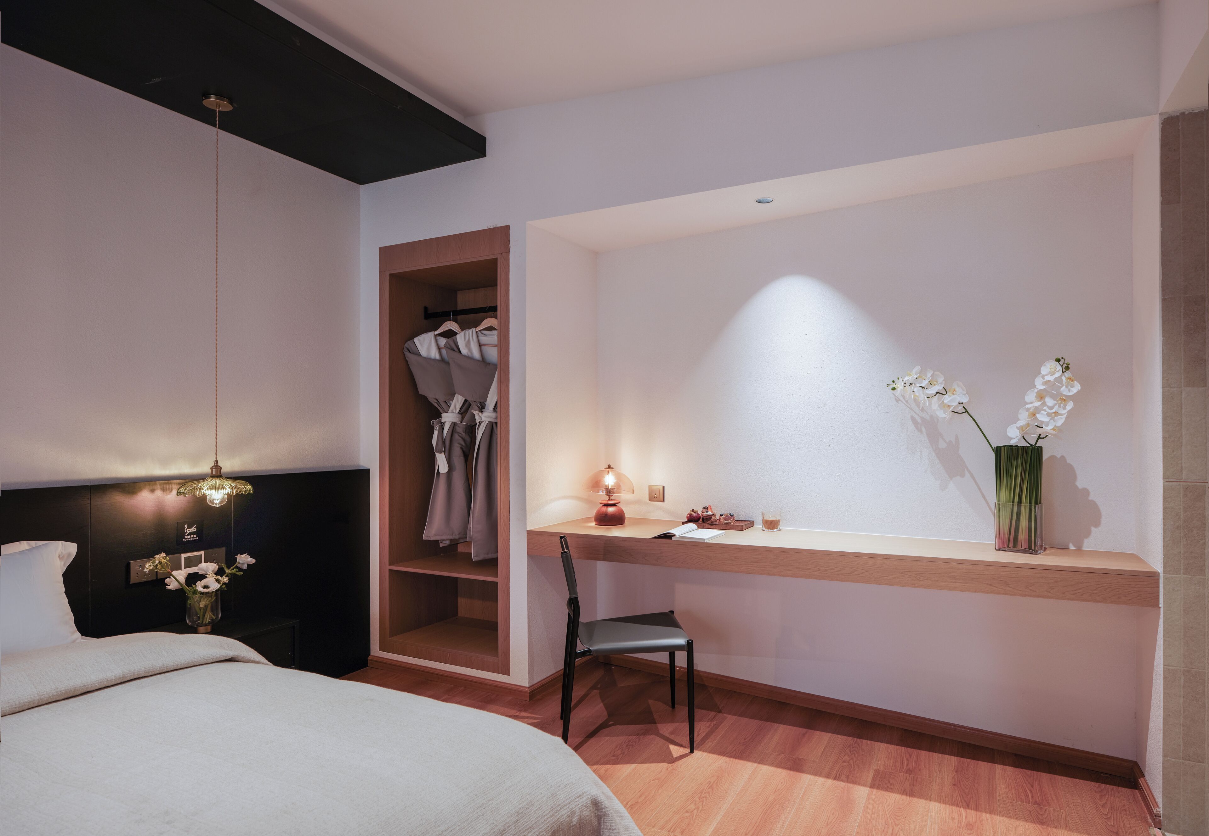 A bedroom with a bed, a desk, a chair, a hanging light, a vase with flowers, and a shelf with clothes.