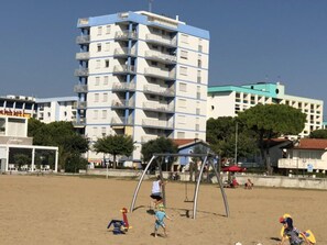 A multi-story building with balconies, a sandy beach area with playground equipment, and a clear sky. - Seafront flat with splendid views from the terrace (Bibione)