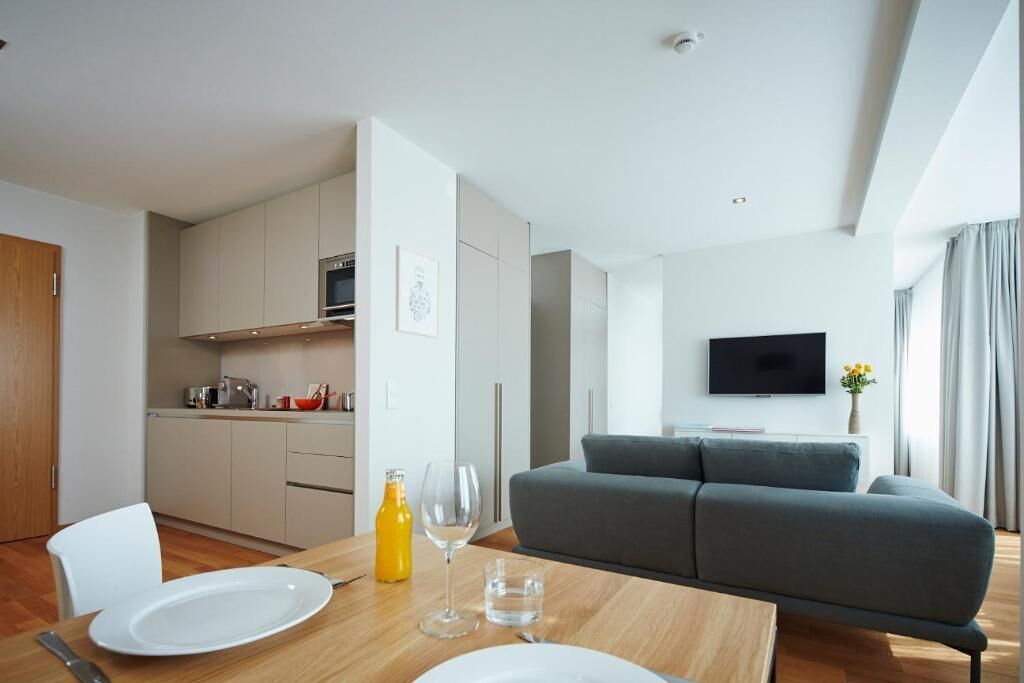 A modern living room with a grey sofa, a dining table with plates and cutlery, a TV mounted on the wall, and a kitchen area with cabinets and appliances.