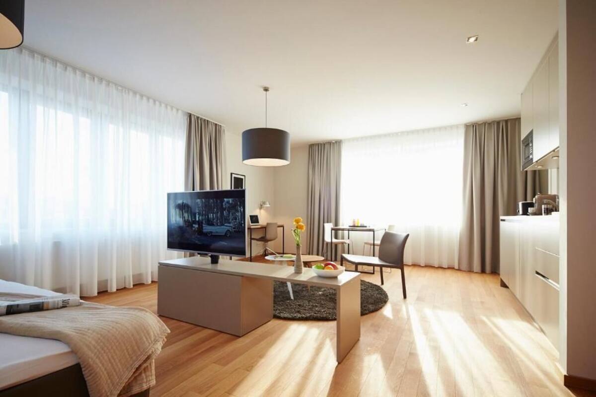 A modern hotel room with a bed, a TV, a dining area, and a kitchen.