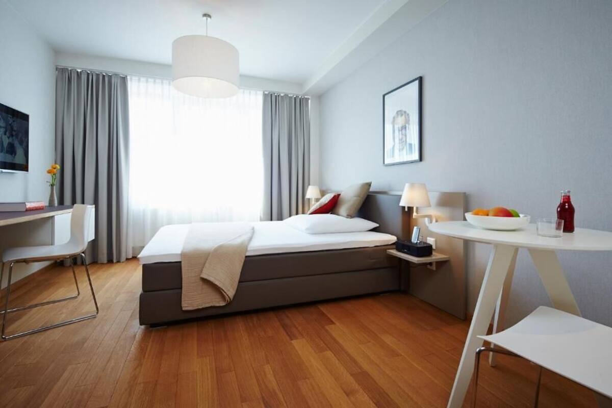 A modern hotel room with a bed, desk, chair, bedside table, and a view of the outdoors.