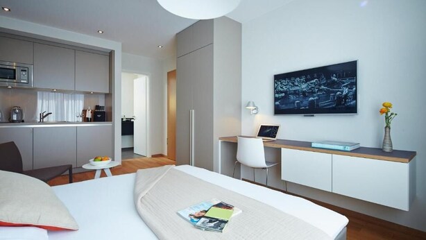 Moderni hotellihuone, jossa on sänky, työpöytä, televisio ja pieni keittiönurkkaus.