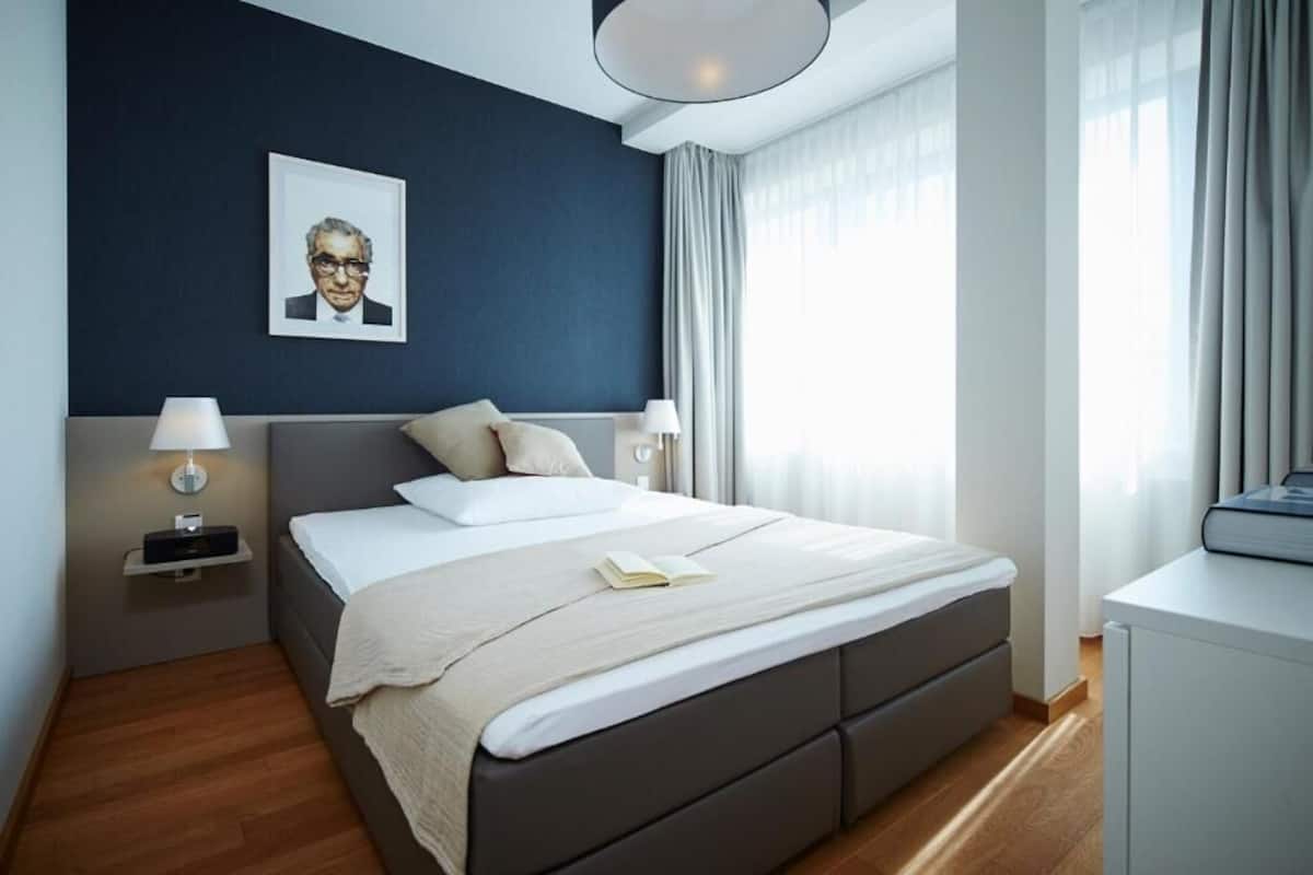 A modern hotel room with a large bed, bedside lamps, a portrait on the wall, and a view of the outdoors through the window.