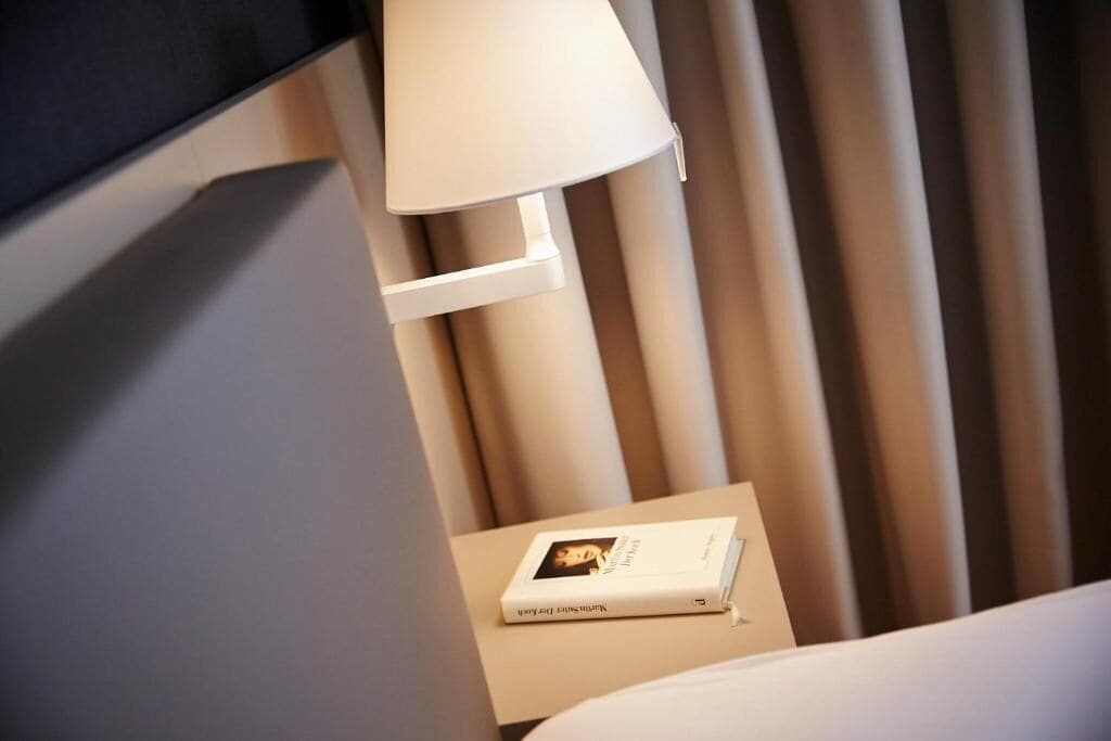 A neatly made bed with a white headboard, a bedside table with a lamp, and a book titled 'The Little Prince' on the table.