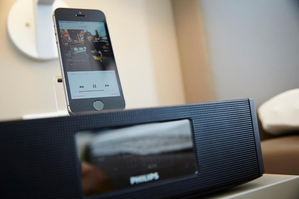 Un smartphone sur une station de chargement, avec une interface de lecteur de musique, un haut-parleur Philips et un lit en arrière-plan.
