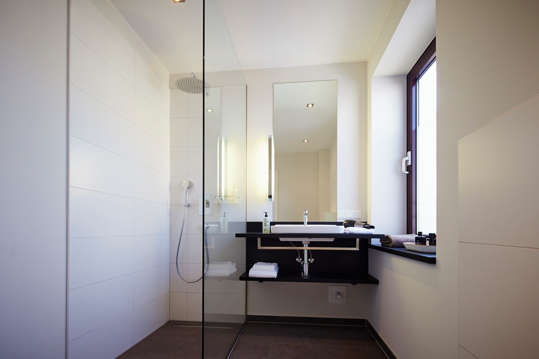 Ein modernes Badezimmer mit Duschkabine aus Glas, Waschbecken und Spiegel sowie einem Fenster.