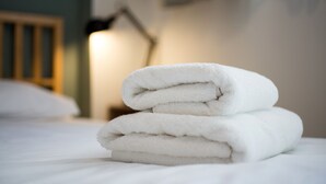 A neatly folded white towel stacked on a bed in a room with a wooden headboard and a lamp in the background.