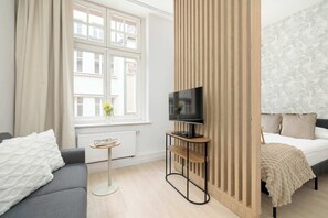 Ein modernes Hotelzimmer mit einer Couch, einem Fernseher auf einem Schrank, einem Bett mit gemustertem Kopfteil und einem Fenster mit Vorhängen.