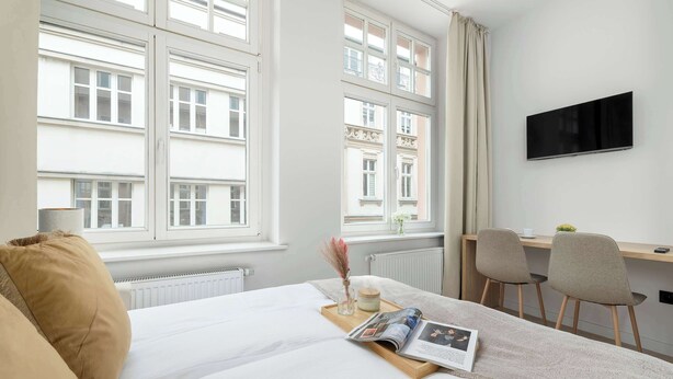 A hotel room with a bed, a desk, a TV, and a window with a view of buildings. - Comfortable Studio in the Heart of Katowice | Remote Work Space (Katowice)