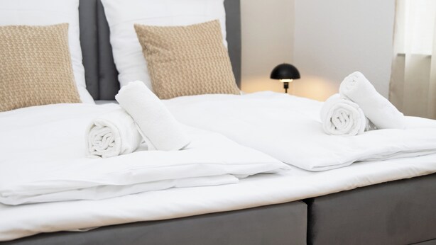 A neatly made bed with white linens and rolled towels, a grey upholstered headboard, and a bedside lamp. - City apartment, incl. parking lot, near Europa-Park & Strasbourg & Black Forest (Offenburg)