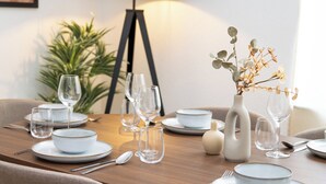 Ett matbord dukat med vita tallrikar, skålar och vinglas, en vas med torkade blommor och en lampa i bakgrunden.