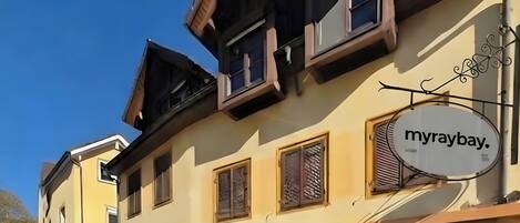 Un bâtiment sur lequel on peut lire l’inscription « myraybay ». Il est doté de nombreuses fenêtres avec volets, d’une antenne parabolique et d’une enseigne métallique décorative.