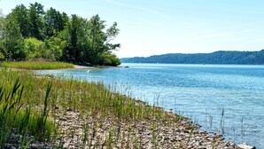 Blick auf einen See mit steinigem Ufer, dichtem Baumbestand und ruhigem Wasser.