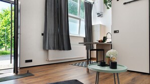 A modern room with a wooden floor, a round glass table, a black ottoman, a radiator, and a window with curtains.