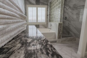 A bathroom with a marble countertop, a bathtub, a shower, and a window with blinds.