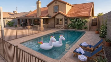 A swimming pool with two swan-shaped floats, a covered patio area with lounge chairs, and a house with a tiled roof.