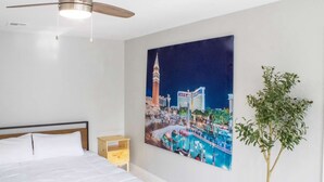 A hotel room with a bed, a ceiling fan, a night sky painting, and a potted plant.