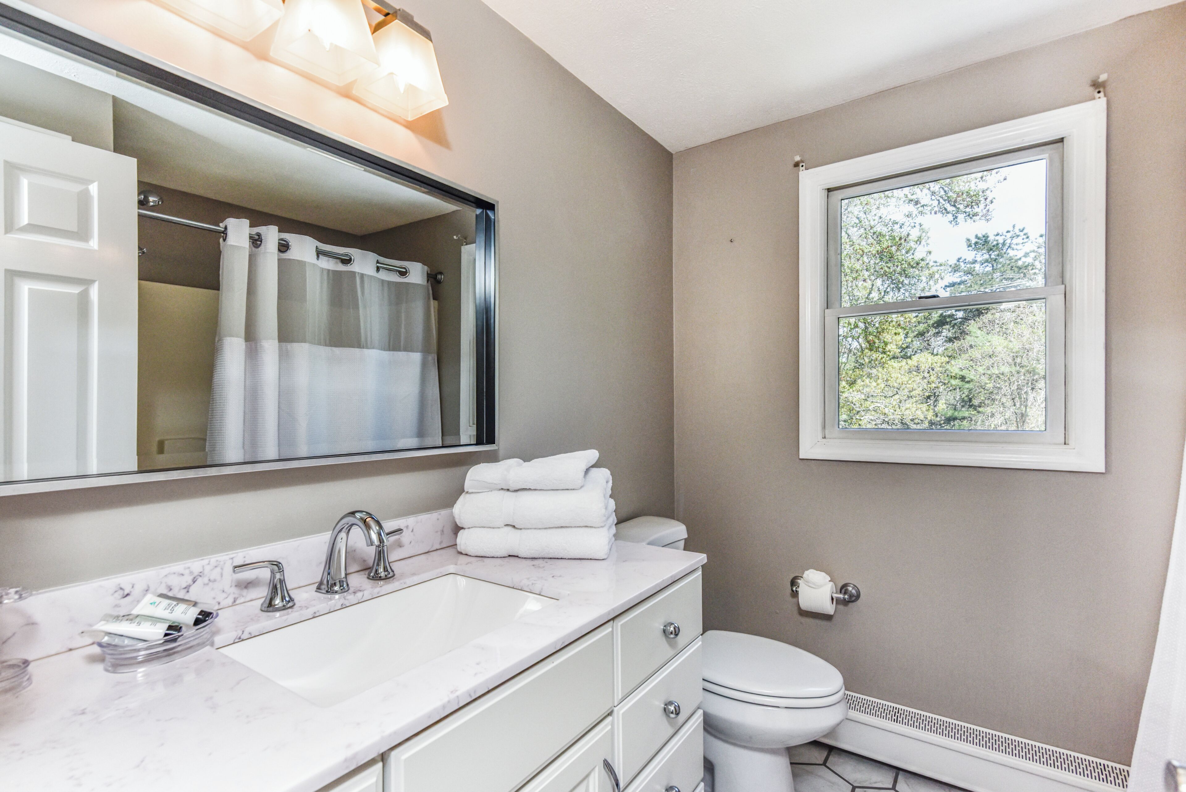 Une salle de bain avec un lavabo blanc, un miroir, des toilettes et une fenêtre donnant sur des arbres.